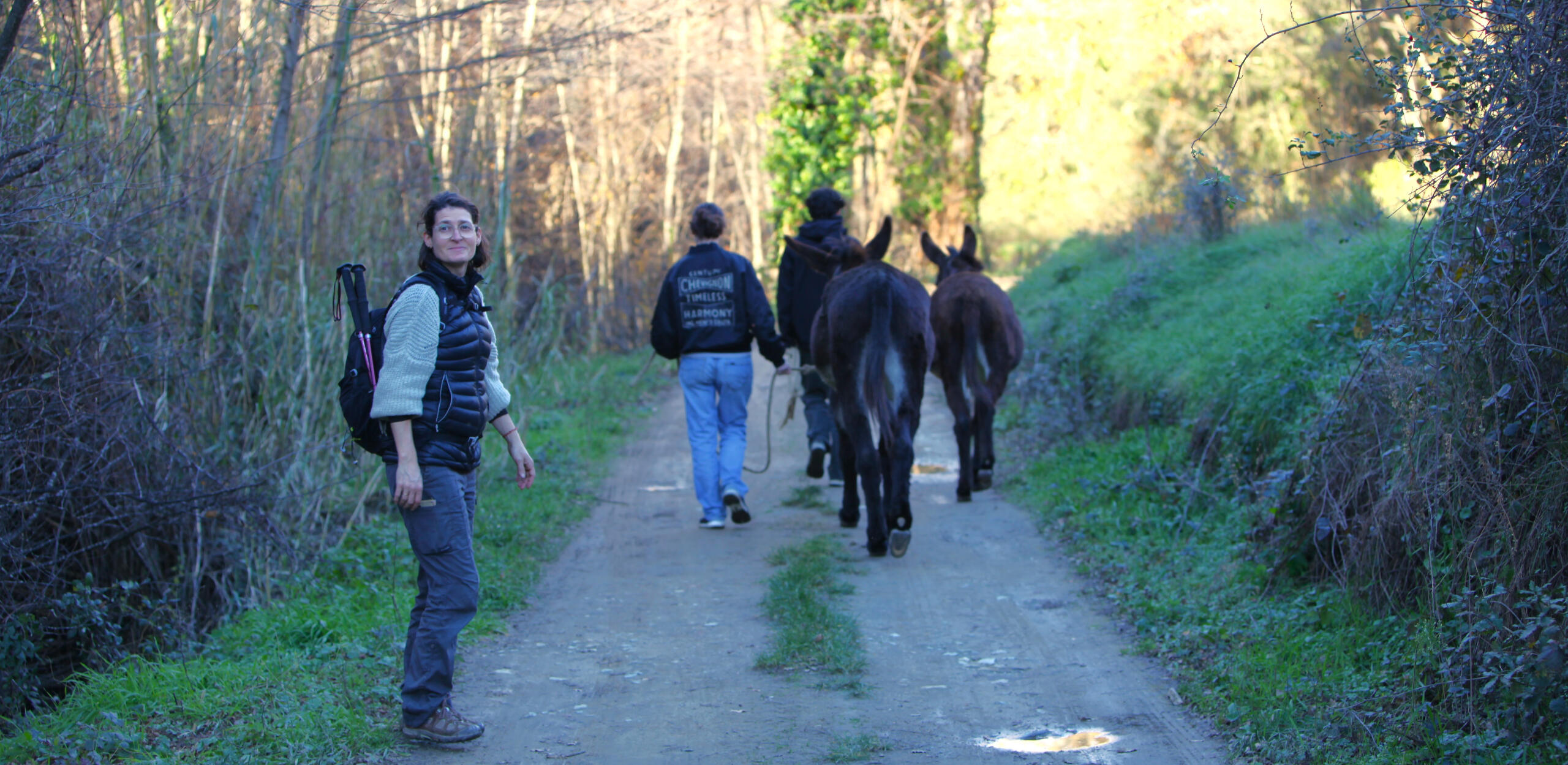 Sur un chemin de campagne, des adolescents accompagnés d'ânes marchent. L'intervenante en médiation animale, se retourne souriante.